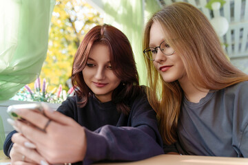 Two fashionable young teenageer completely absorbed in the screen of a smartphone casually unwind at an alfresco summer cafe.