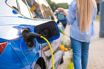 An electric vehicle or electric car is connected to a charging station as a person walks away holding groceries.