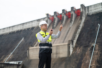 Smiling young Asian civil engineer standing with his arms crossed wearing a helmet and using a walkie-talkie. View and inspect the operation of the dam's sluice gates using the Quality Monitor tablet.