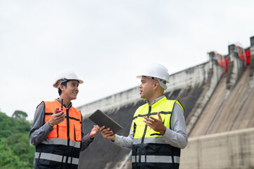 Smiling young Asian civil engineer standing with his arms crossed wearing a helmet and using a walkie-talkie. View and inspect the operation of the dam's sluice gates using the Quality Monitor tablet.