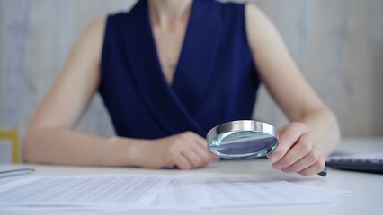 Fototapeta premium Businesswoman holding magnifying glass analyzing financial documents at office desk. Taxes, audit in business