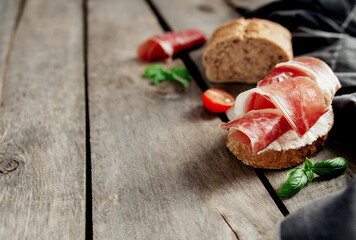 Italian cuisine concept. Crusty bread toast with cream cheese and thinly sliced ham, cherry tomatos, basil leaves branches, kitchen towel on wooden background. Lunch in Italy, healthy tasty break 