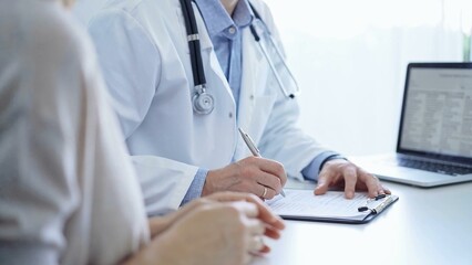 A doctor and a patient. The physician, wearing a white medical coat over a blue shirt, is filling out a medical record form during a consultation in the clinic. Medical service