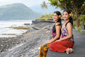 Two Balinese women in traditional attire sitting by the seaside