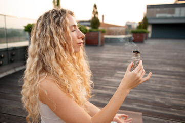 Woman with long curly hair holds a smudge stick, releasing smoke, on a rooftop deck during sunset....