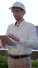 Man an architectural engineer, wearing a white shirt and hard hat, is using a digital tablet while inspecting a building site at sunrise, front and vertical view