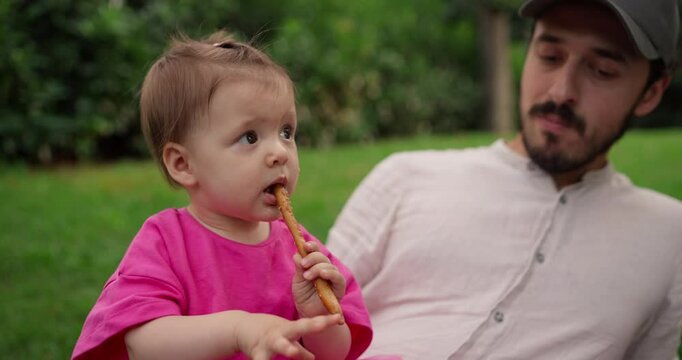 Little girl in pink dress chewing breadstick while relaxing with her dad in park on lawn in summer. Little girl in pink dress sits in her dad arms and relaxes with her family on a mat on the lawn in