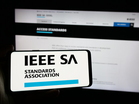Stuttgart, Germany - 10-07-2024: Person holding cellphone with logo of IEEE Standards Association on screen in front of webpage. Focus on phone display.