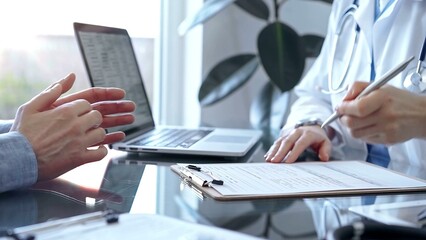 Doctor woman wearing white medical coat is consulting patient in clinic office while both are sitting at the glass desk. Physician is listening and making notes. Medicine concept