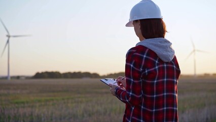 Adult woman engineer wearing white cask is taking notes on a clipboard on a field with wind turbines, as the sun sets, back view