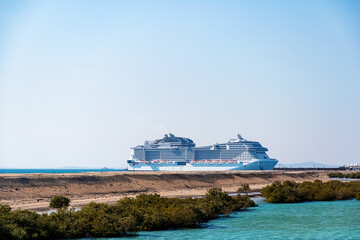 A large cruise ship is docked at a island port, with clear blue skies above. Ship is surrounded by a sandy beach and lush greenery.