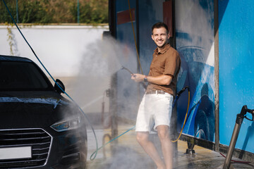 Happy young man at a car wash uses a pressure washer and sprays water uphill. Man is having fun