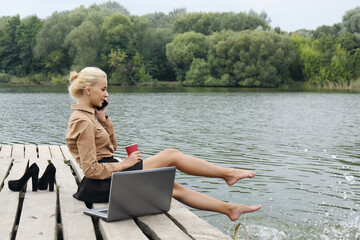 Woman sitting on a pier by the river drinking coffee, talking on the phone and splashes water with his feet near the laptop