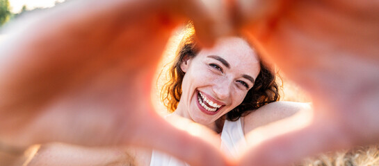 Smiling woman making a heart shape with hands - Pretty joyful woman laughing at camera outside -...