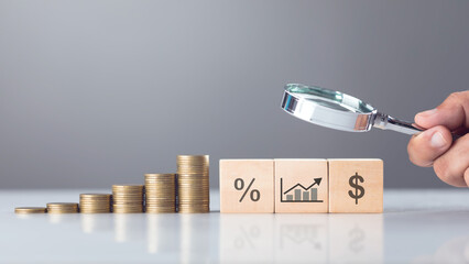 Wooden block labeled interest icons sits beside a stack of coins, while a hand holds a magnifying glass over them. The image symbolizes the scrutiny of taxes and financial analysis