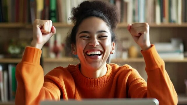 A woman in an orange sweater is smiling and holding her hands up in the air. She is looking at a laptop computer