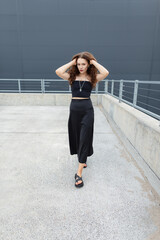 Stylish beautiful urban fashion woman in black top with skirt and sandals walking in city against background of building