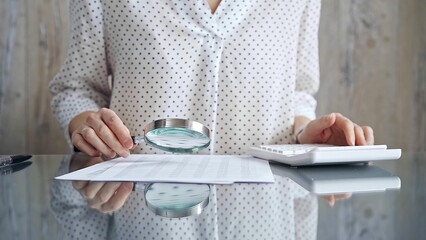 Auditor woman analyzing financial documents with magnifying glass and calculator at her office desk in formal white blouse with polka dots. Business people concept
