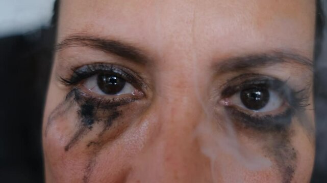 woman, whose make-up has runny from crying, smokes a cigarette and looks through the smoke.