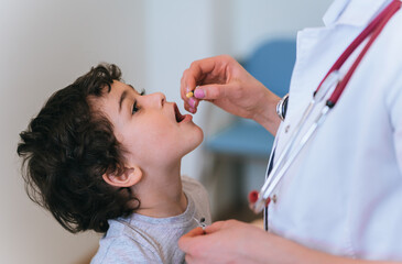 A healthcare professional gives medicine to a young boy, who looks up with trust and curiosity in a bright, clinical setting.
