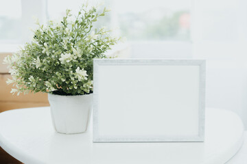 A blank picture frame beside a lush green plant on a bright, airy table in a sunlit room