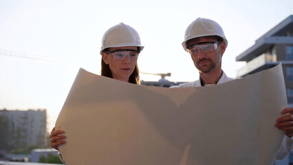 Two architects colleagues, wearing safety helmets and glasses, are examining blueprints at a construction site during sunrise or sunset, front view