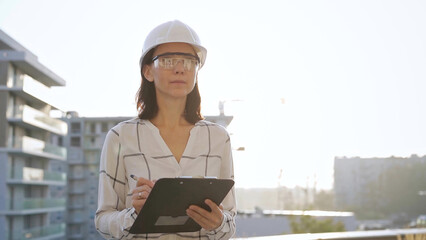 Woman construction engineer wearing white checked blouse and hard hat is making notes on a clipboard while inspecting a building site at sunset, front view. Architecture and engineering concepts