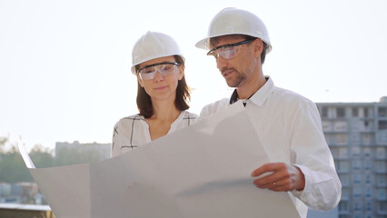 Two architects, wearing safety helmets and glasses, are examining blueprints at a construction site during sunrise or sunset, front view. Architecture and engineering concepts