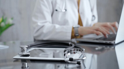 Close-up of doctor's desk with stethoscope and tablet computer. Physician is using a laptop at the background. Medicine concept