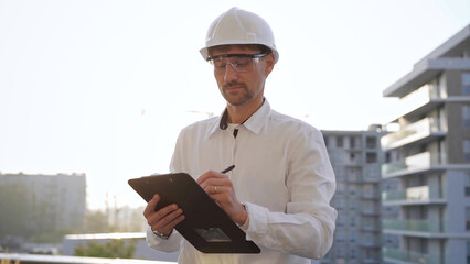 Man construction engineer wearing white shirt and hard hat is making notes on a clipboard while inspecting a building site at sunset, front view. Architecture and engineering concepts