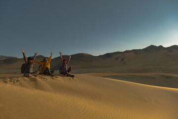 A group of people are sitting on the sand during the golden hour. Friends sit and raise their hands on the sand during sunset. A golden hour in the desert with people. Travelers are friends. Happy peo