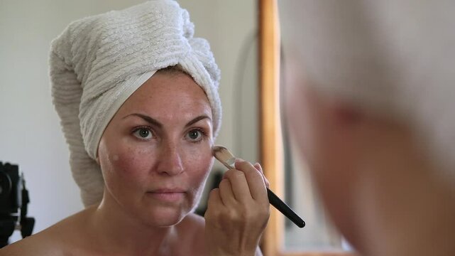 Woman does makeup in front of a mirror applies foundation and concealer with brush, after showering with a towel on her head