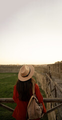 Back view of a girl tourist in a hat with a backpack standing at sunset in an ancient fortress looking at the sky