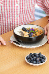 A woman is sitting at a table, eating a healthy breakfast. Granola with Greek yogurt and blueberries