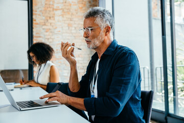 Mature businessman speaking on phone while using laptop in modern office setting with colleague...