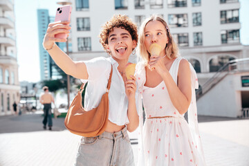 Two young beautiful smiling hipster female in trendy summer clothes. Carefree women posing on street background. Positive models eating tasty ice cream in waffles cone in sunny day, take selfie photo