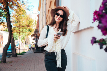 Young beautiful smiling woman in trendy summer clothes. Carefree female posing in street in sunny day. Positive model holds smartphone, looks at cellphone screen, uses mobile apps, in hat, eyewear