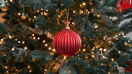 Christmas red ornament close-up Christmas tree flickering light hanging on beautifully lit. Festive bauble is surrounded by glowing string lights pine branches, creating warm cozy holiday atmosphere.