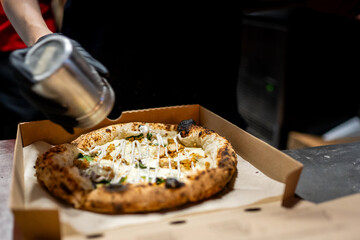 A close-up of a hand sprinkling seasoning on a freshly baked pizza in an open cardboard box. The pizza is topped with various ingredients, making it look delicious and ready to eat