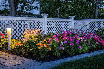 Obraz premium A cozy terrace showcases a beautiful flower bed at night, with a white ground lamp casting a gentle glow on the colorful flowers and shrubs. The low white fence encircling the garden adds charm and