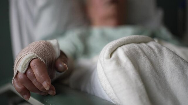 A close-up view of a hand bandaged in a hospital bed, with a blurred background of a patient's chest and white sheets.