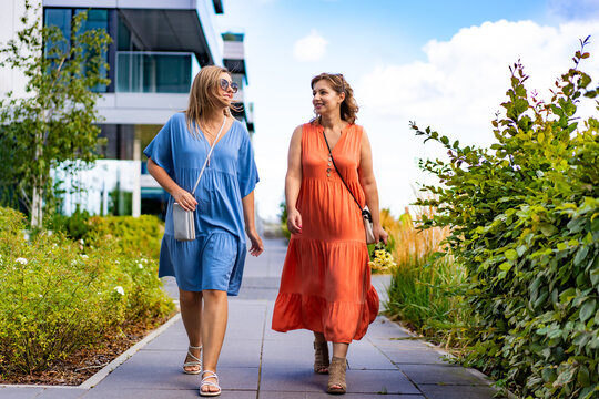Summer in city. Two young beautiful women wearing light dresses, bags and sunglasses, talking while walking around modern housing estate in city on sunny day. Meeting of friends.	