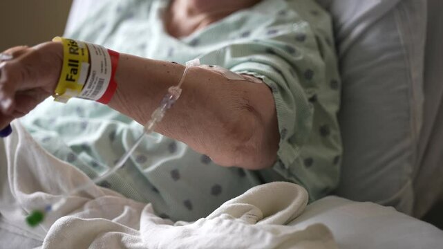 A close-up shot of a patient's arm in a hospital bed, wearing a yellow 'Fall Risk' bracelet and an IV line. The patient is wearing a green and white striped gown and is lying under a white blanket.