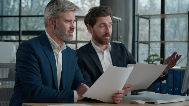 Two Caucasian men businessmen in office working together on financial statistics analyzing charts project discussing data in report doing paperwork using computer looking documents client consultation