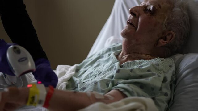An elderly woman lies in a hospital bed, wearing a hospital gown and an ID bracelet. A medical professional is scanning her wrist with a device. The woman has a black eye and recovering from injury.