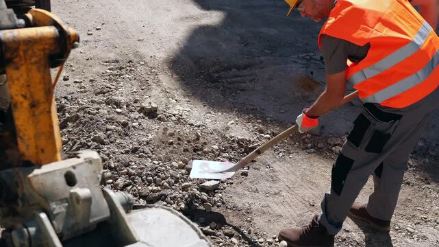 A construction worker in safety gear is actively repairing a road surface, digging with a shovel under bright sunlight in a bustling urban environment.