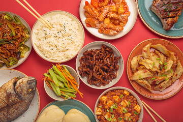 Variety of delicious chinese dishes on plates and bowls arranged on red background: kung pal chicken, sweet and sour pork, noodles, dumplings, mapo tofu, fish, rice top view