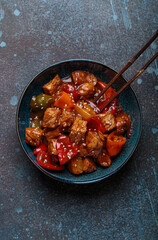 Sweet and sour pork Chinese dish with bell peppers and pineapple sprinkled with sesame seeds, served in a blue bowl with chopsticks on a dark background