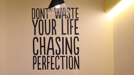 Lightbulb Illuminating over a Wall with a Quote Saying Don't Waste Your Life Chasing Perfection,...