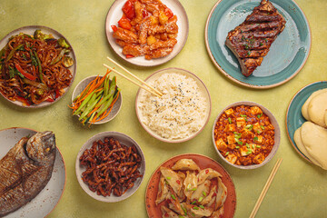 Variety of delicious chinese dishes on plates and bowls arranged on green background: kung pal chicken, sweet and sour pork, noodles, dumplings, mapo tofu, fish, rice top view with copy space